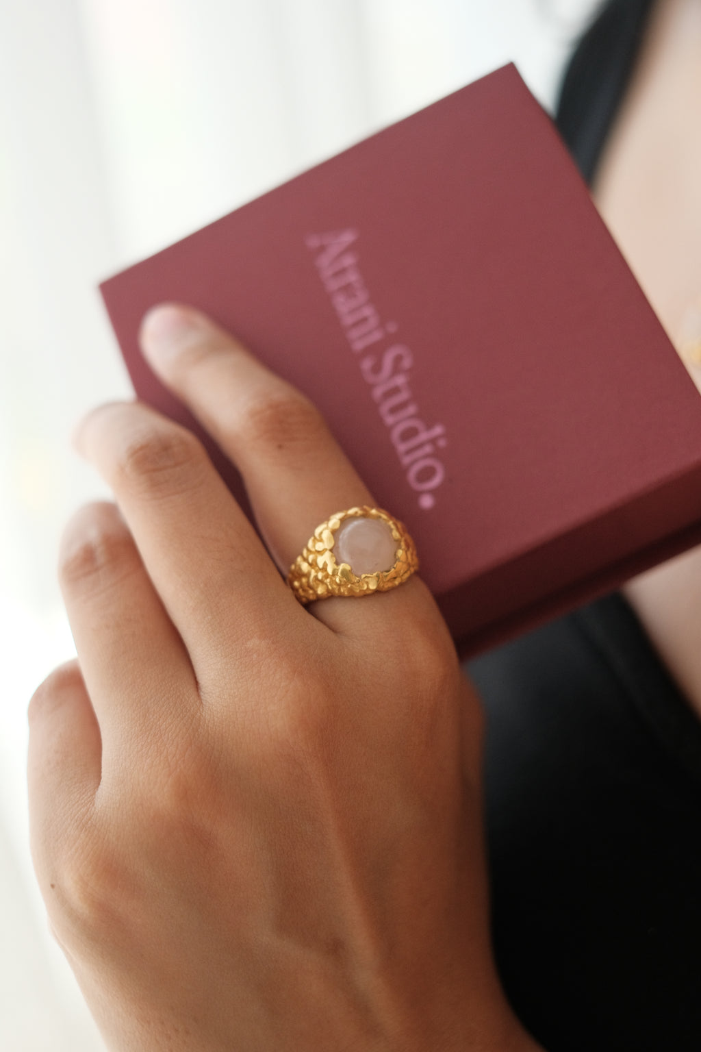 Hand wearing a gold ring with a pink stone on a red box with 'Atrani Studio' branding.